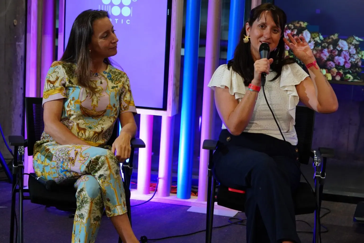 Dos mujeres conversan sentadas en un escenario del CRTIC, una con micrófono, bajo luz azul y violeta y con la pantalla del centro detrás.