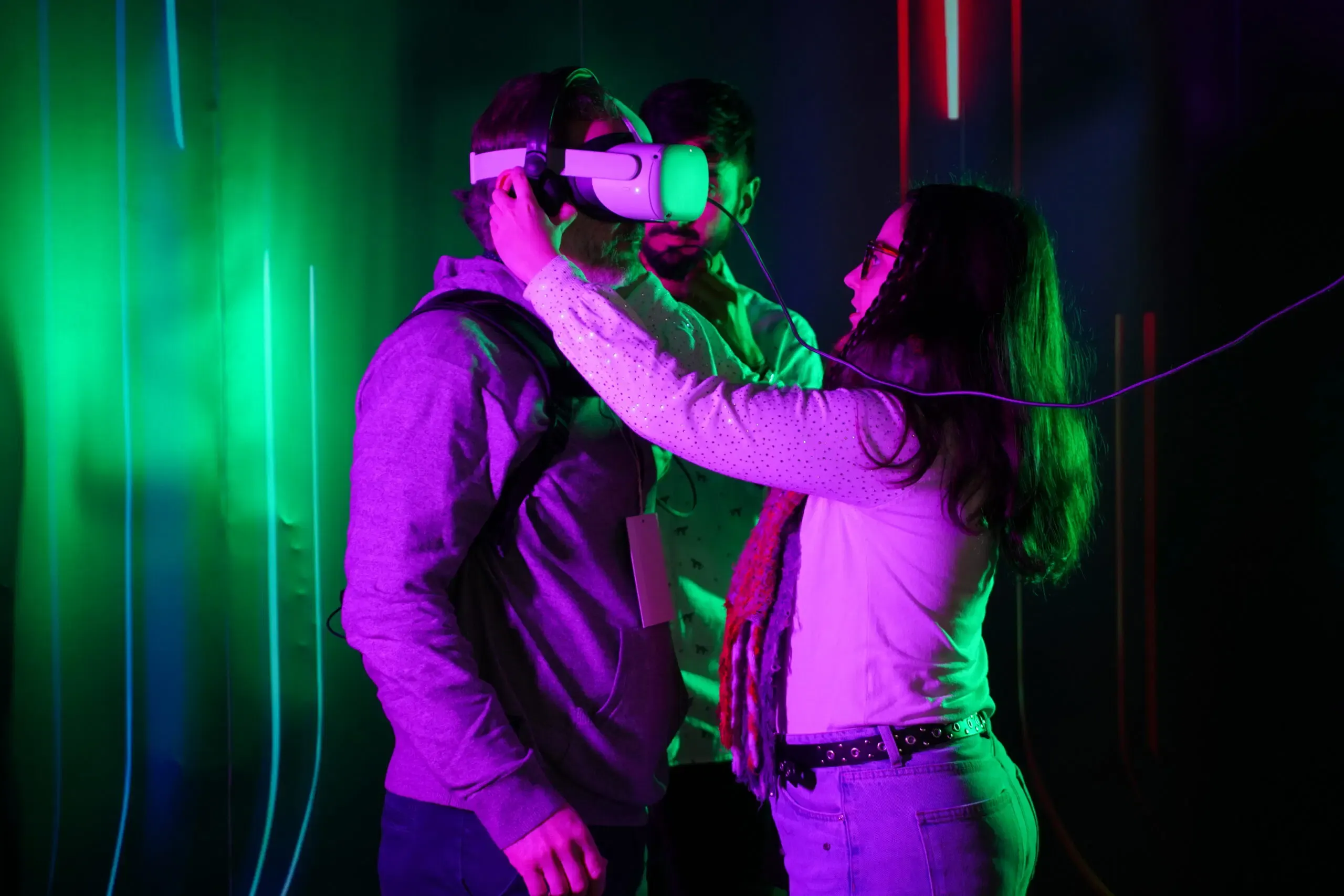 Una instructora ajusta unas gafas de realidad virtual a un participante en el laboratorio, iluminados por tubos de neón verde y magenta.